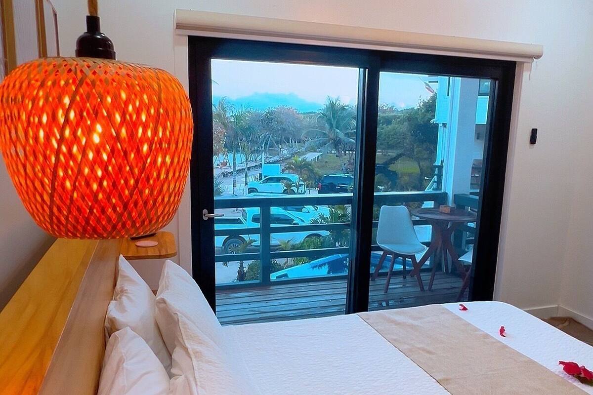 A bedroom with a bed, white pillows, a wooden bedside table, a red patterned lamp, and a view of a balcony with outdoor furniture.