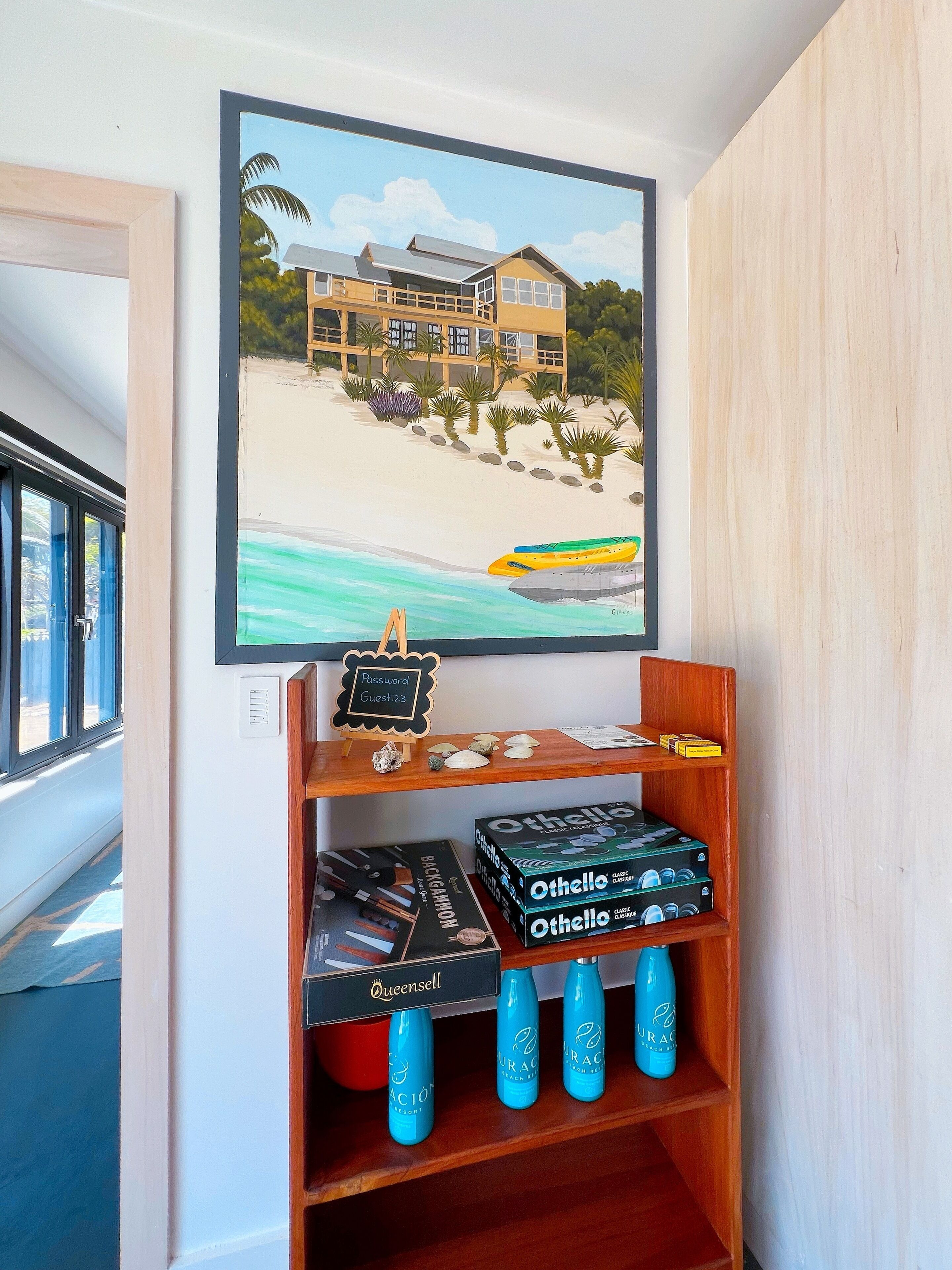 A framed painting of a beach house, a wooden shelf with board games and bottled water, and a sign with a password and guest number.