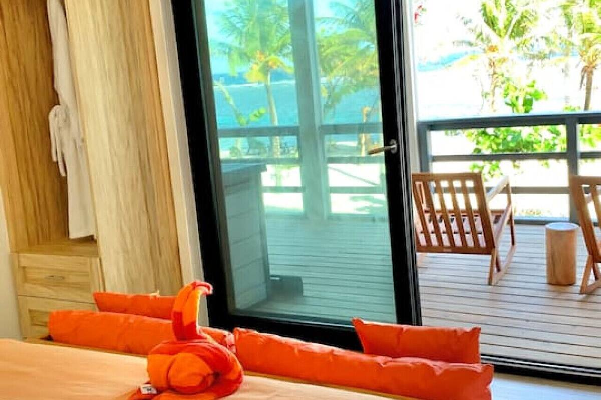 A bedroom with a bed, orange pillows, a wooden headboard, a ceiling fan, and a sliding glass door leading to an outdoor deck with chairs.