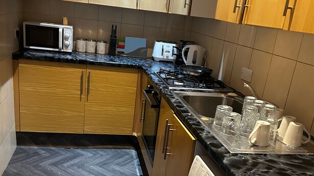 A compact kitchen with wooden cabinets, a microwave, a toaster, and a washing machine. - Stunning 4bedrooms Flat (Manchester)