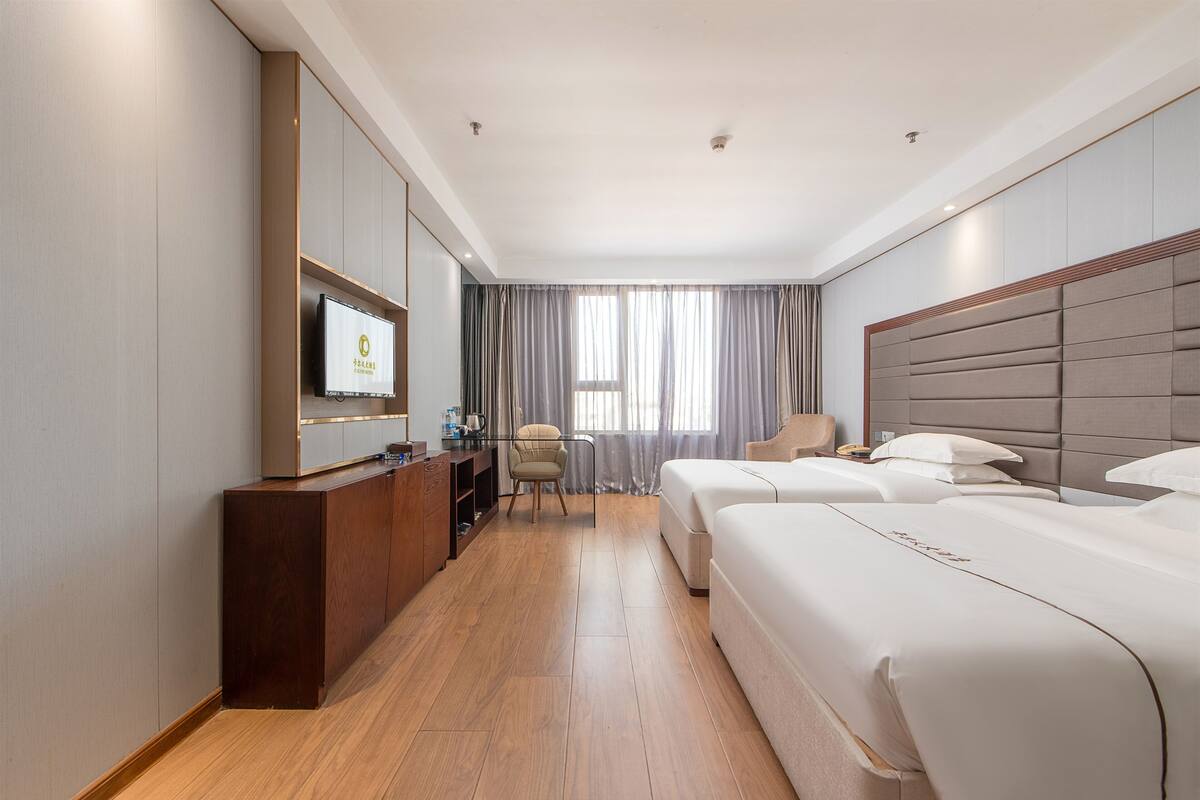 Habitación de hotel con suelo de madera, un escritorio con televisor, dos camas, una silla y una ventana con cortinas.