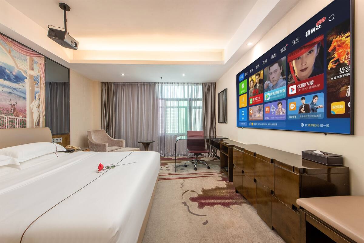 Habitación de hotel con una cama grande, un escritorio, una silla, un televisor montado en la pared mostrando varios canales y una ventana con cortinas.