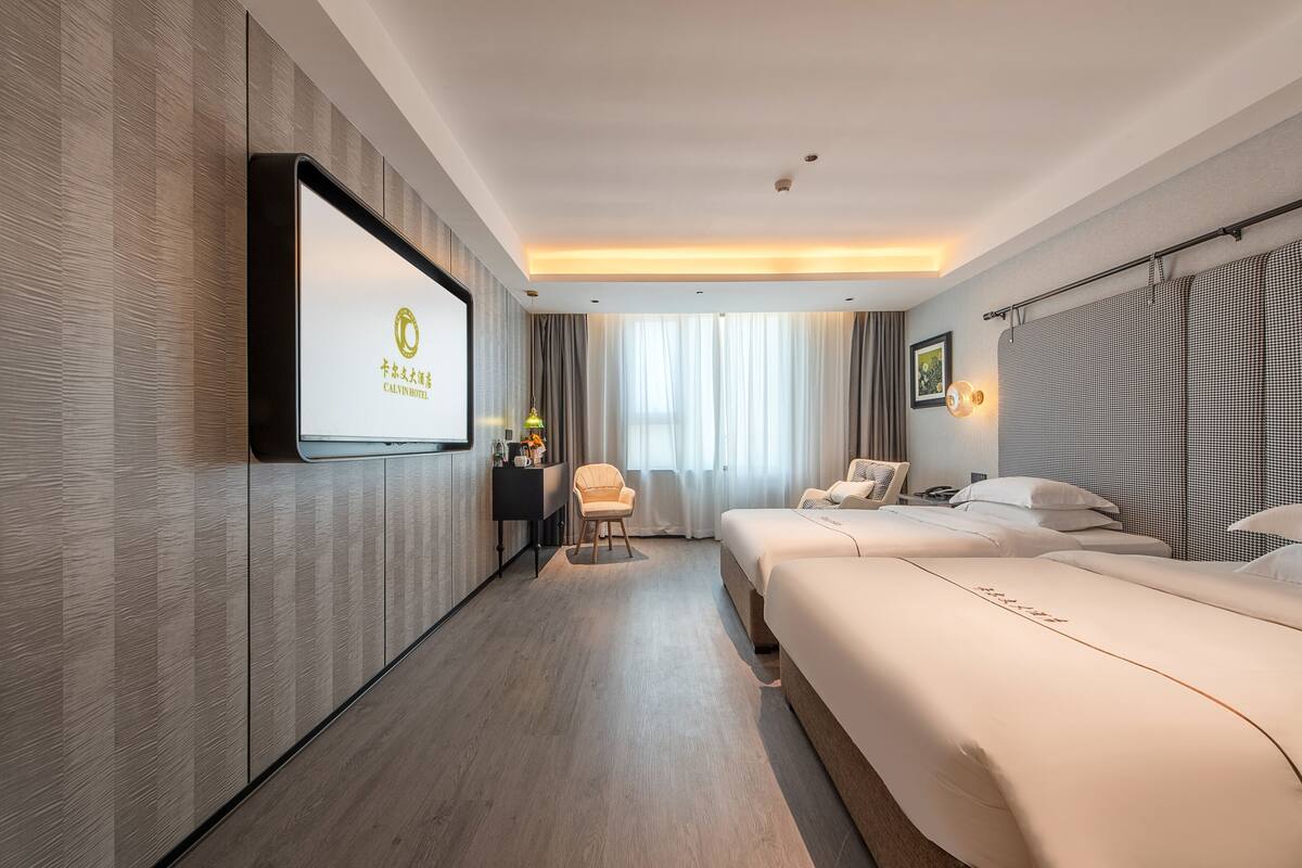 Habitación de hotel con una cama grande, una cama más pequeña, un televisor montado en la pared, un escritorio con una silla y una ventana con cortinas.