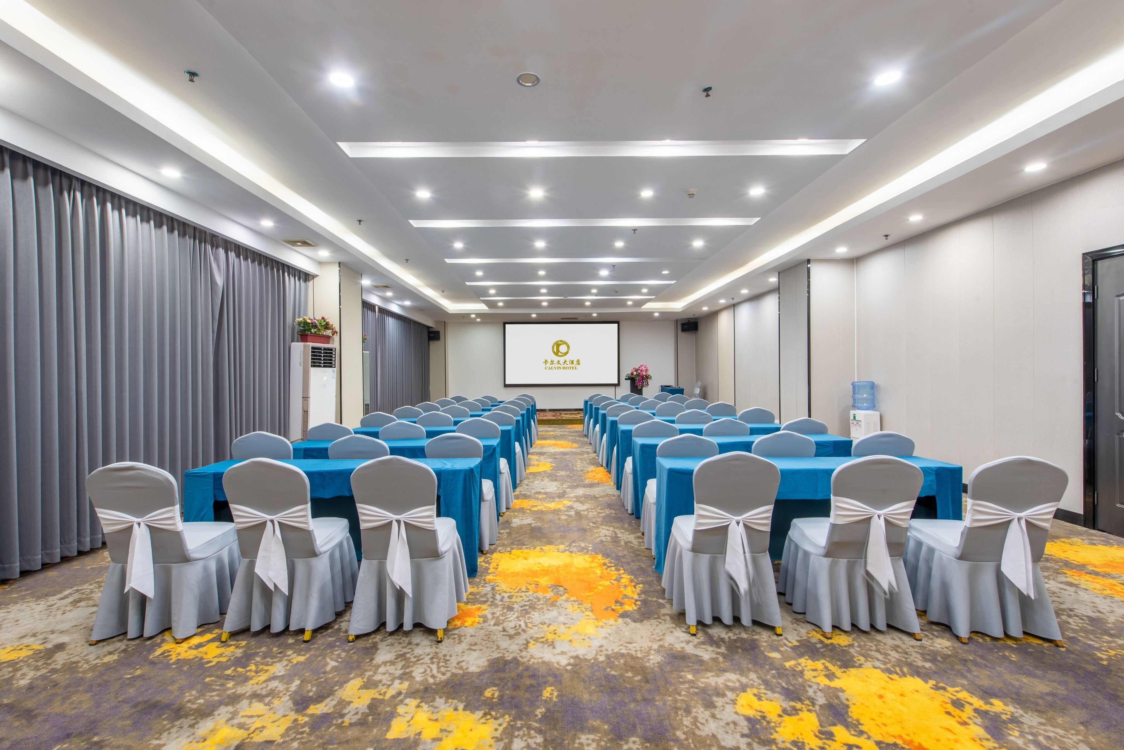 A conference room with rows of tables and chairs, a projector screen, and a water dispenser.