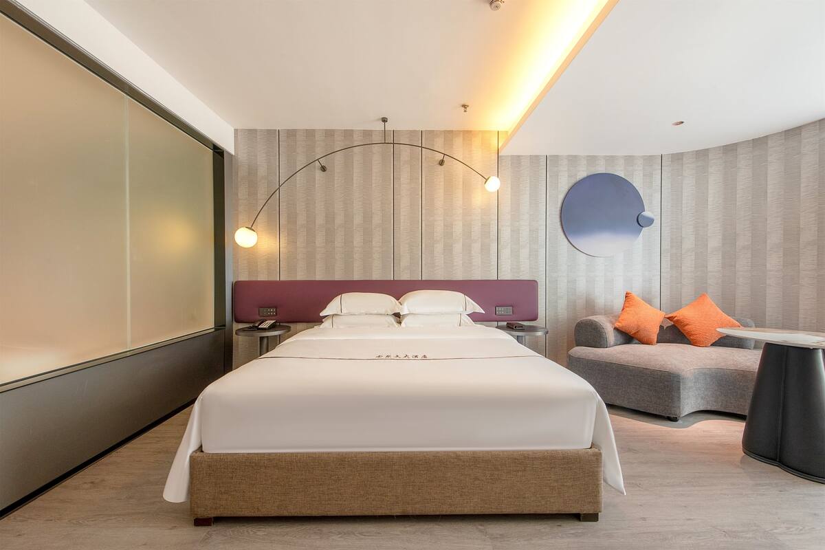 Una habitación de hotel moderna con una cama grande, un sofá, un espejo redondo y una mesita de noche.