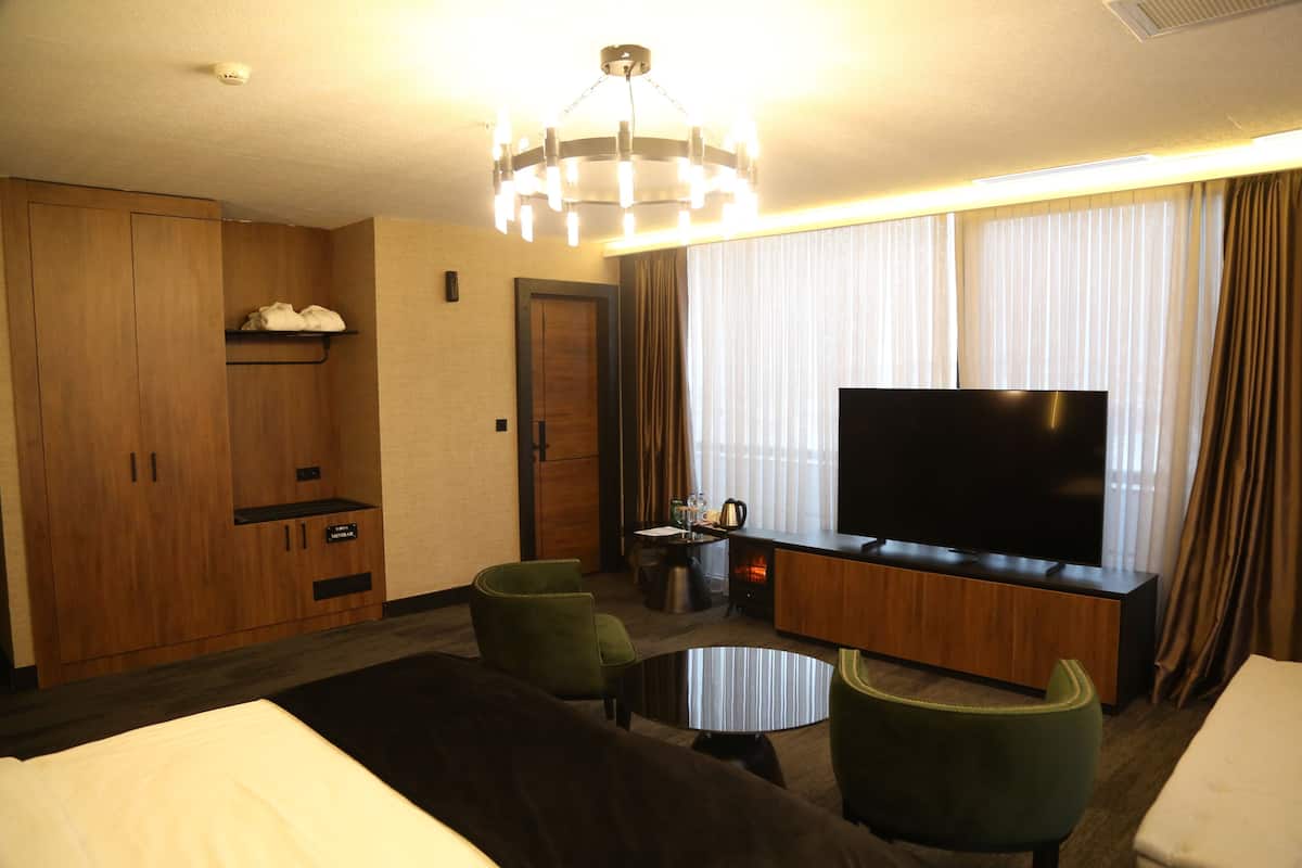 A hotel room with a large flat-screen TV, a sofa, two armchairs, a coffee table, a minibar, and a wooden wardrobe.