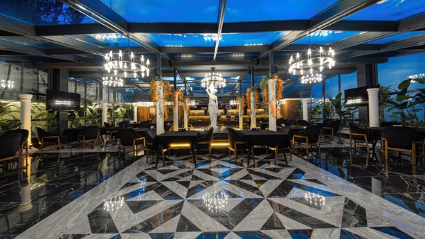 Un vaste espace de restauration intérieur doté d’un sol en marbre à motifs géométriques, de lustres modernes et offrant une vue sur le ciel grâce à un plafond en verre.