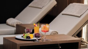 Une pièce comprenant deux fauteuils inclinables, une table d’appoint en bois sur laquelle sont posés un plateau de fruits et deux boissons, ainsi qu’une fenêtre laissant entrer la lumière naturelle.