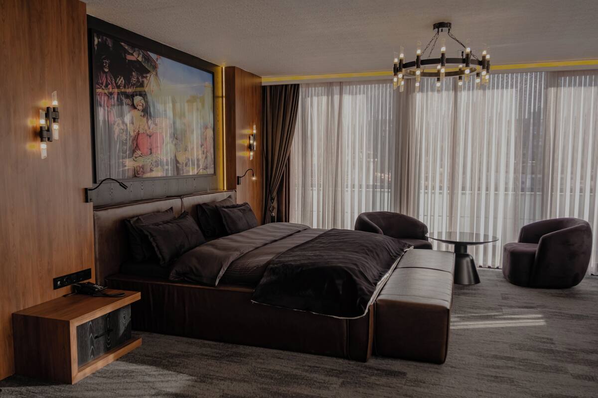 A modern hotel room with a large bed, a small table, two armchairs, a chandelier, and a large painting on the wall.