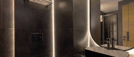 A modern bathroom with a dark tiled wall, a toilet, a sink with a marble countertop, and a large mirror.