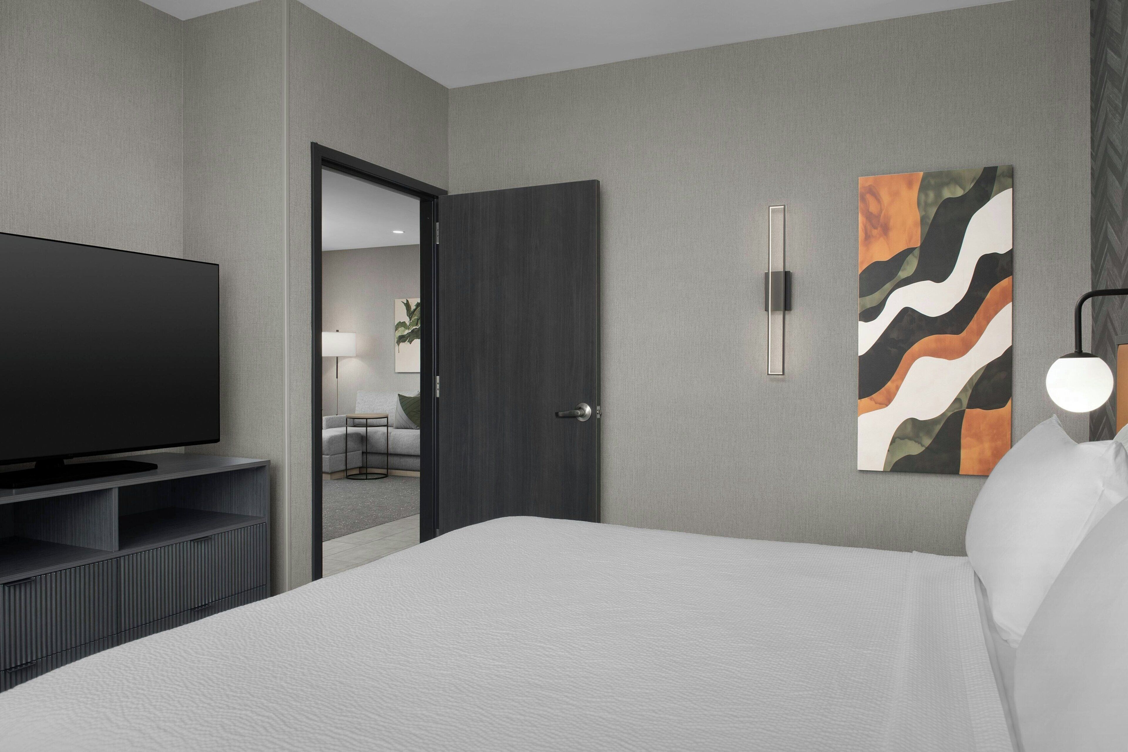 A modern hotel room with a large flat-screen TV, a bed with white linens, a dark wooden door, and a wall-mounted abstract art piece.