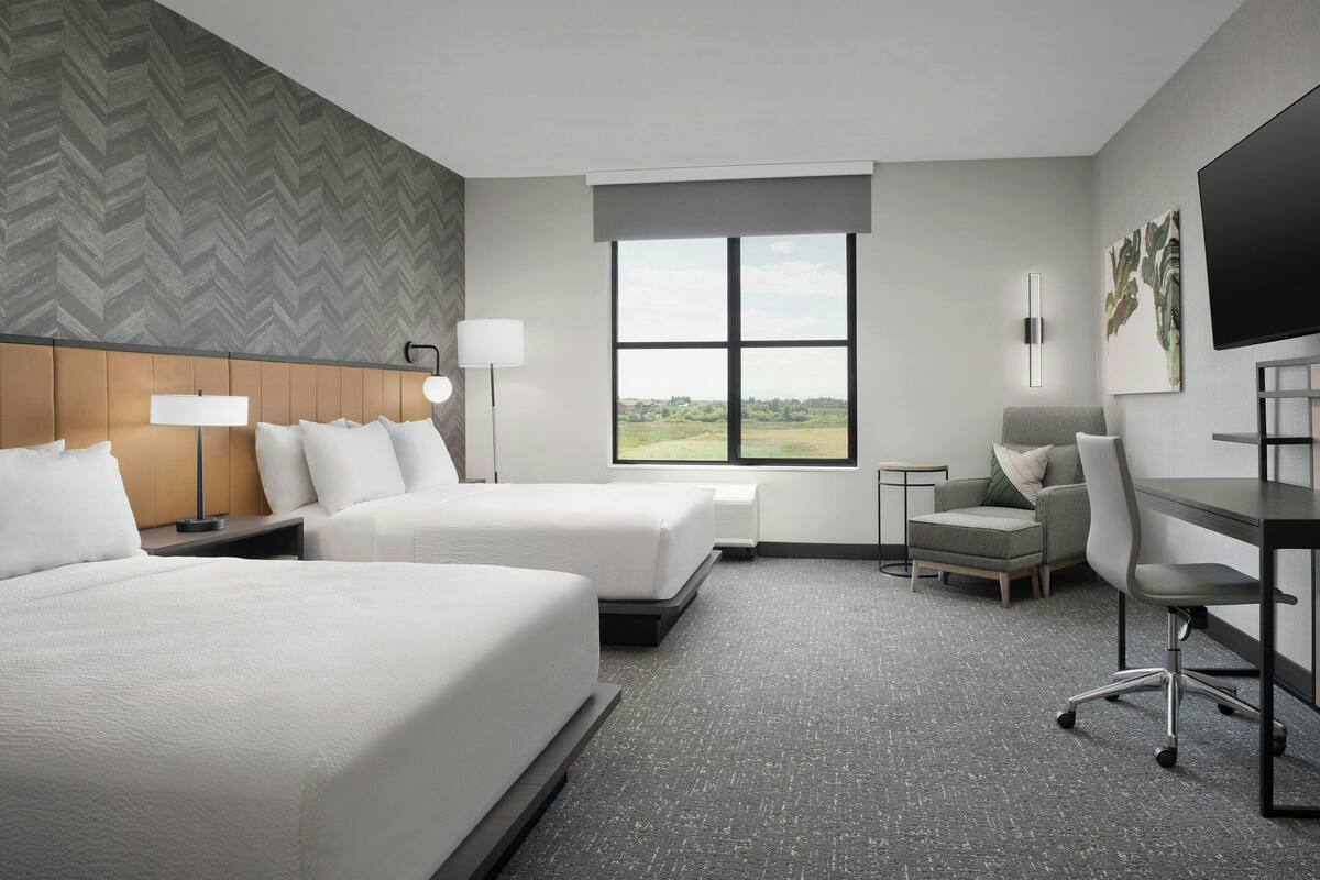 A hotel room with two beds, a desk, a chair, a TV, and a window with a view of the countryside.