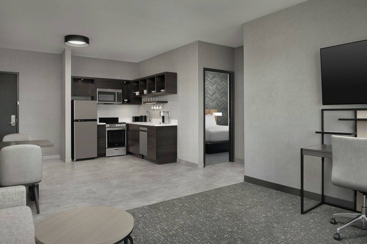 A modern hotel room with a kitchenette, a flat-screen TV, a sofa, and a dining table.
