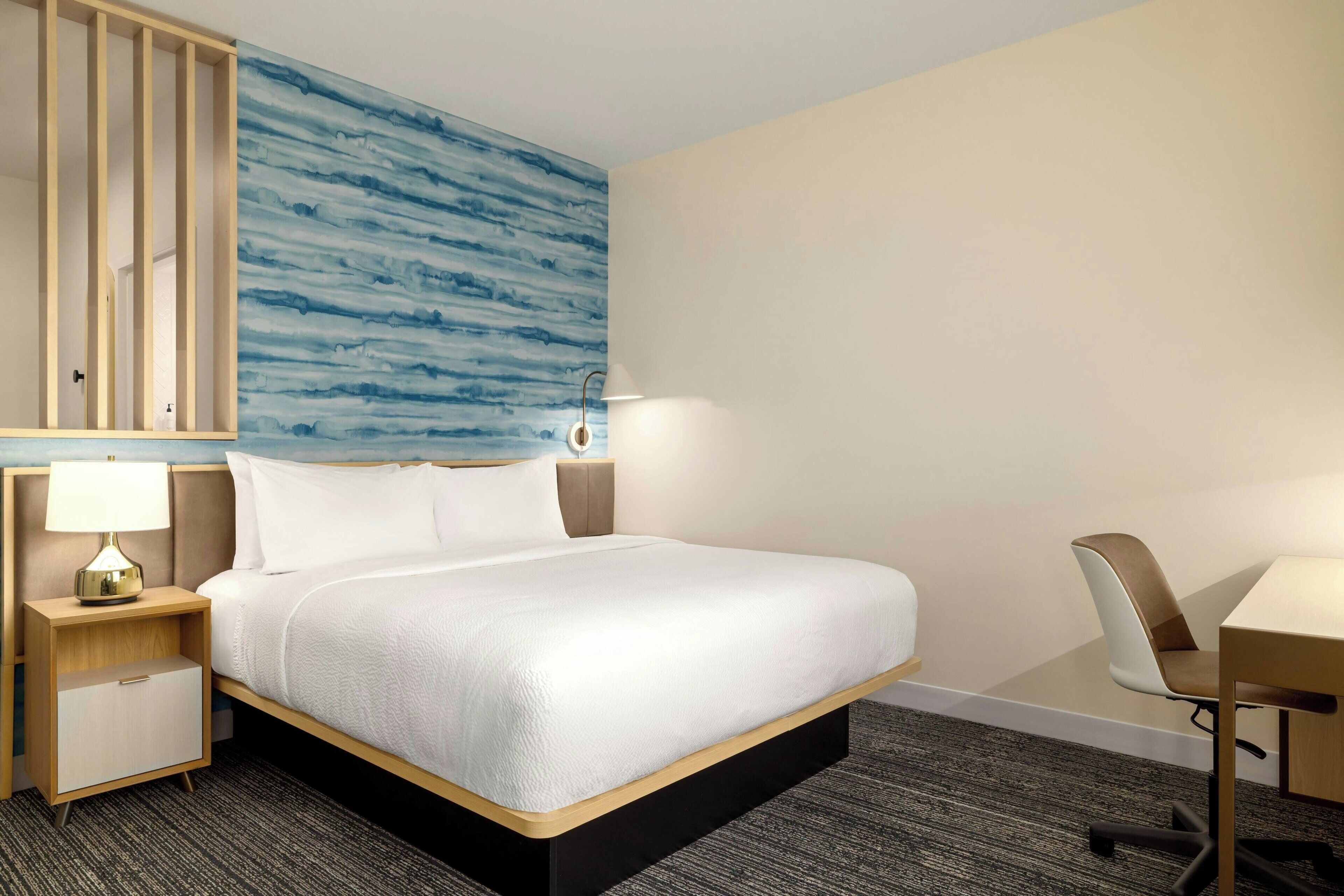 Une chambre d’hôtel comprenant un lit, un bureau, une chaise, une table de chevet avec une lampe, et un mur orné d’un motif abstrait bleu.