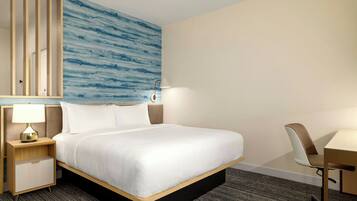 A hotel room with a bed, a desk, a chair, a nightstand with a lamp, and a wall with a blue abstract design.