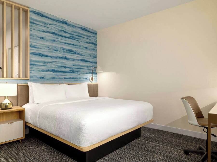 A hotel room with a bed, a desk, a chair, a nightstand with a lamp, and a wall with a blue abstract design.