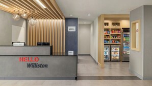 A reception desk with a 'HELLO Williston' sign, a kitchenette area with a refrigerator and shelves stocked with snacks and beverages, and a wall with a 'Marriott Bonvoy' sign. - TownePlace Suites by Marriott Williston (Williston)