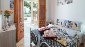 A bedroom with a bed, a wooden door, a small table with a vase, and a painting on the wall.