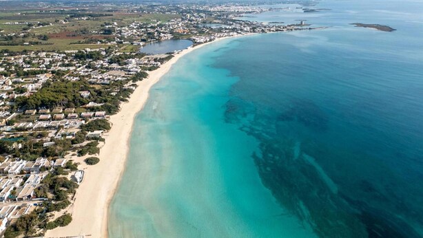 An aerial view of a coastal area with a long sandy beach, a clear blue sea, and a densely populated residential area with houses and buildings.