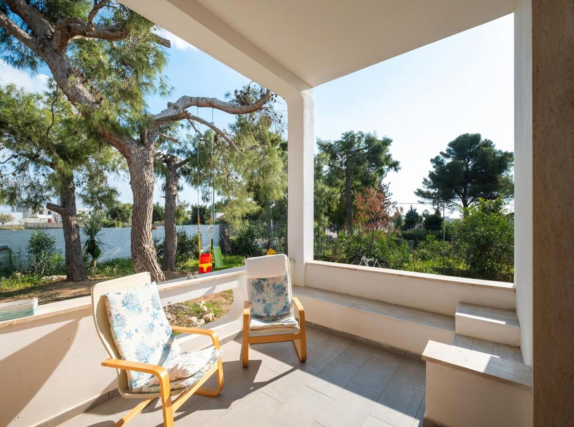 A balcony with two wooden chairs and a bench, overlooking a garden with trees and a swing.