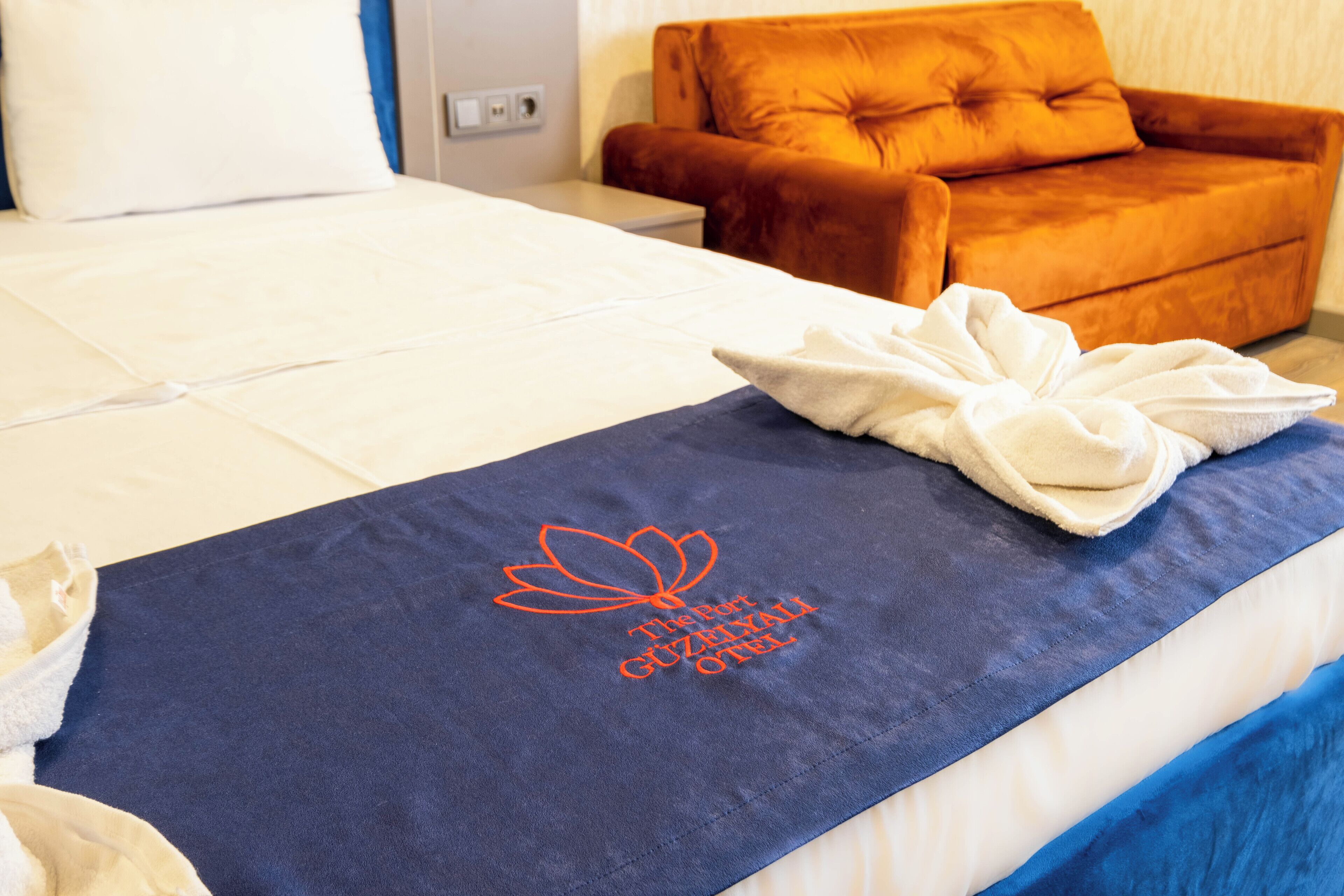 A hotel room with a neatly made bed, a blue towel with embroidered text, and an orange armchair.