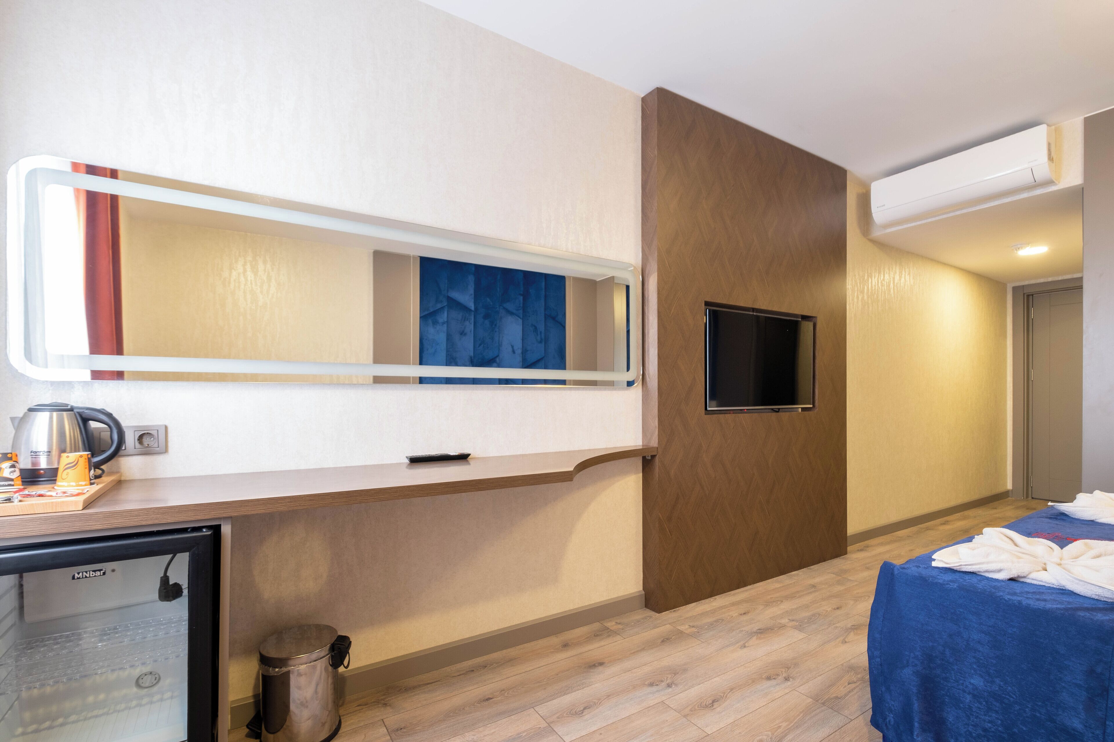 A hotel room with a minibar, a television, a mirror, a bed with a blue cover, and a wooden floor.