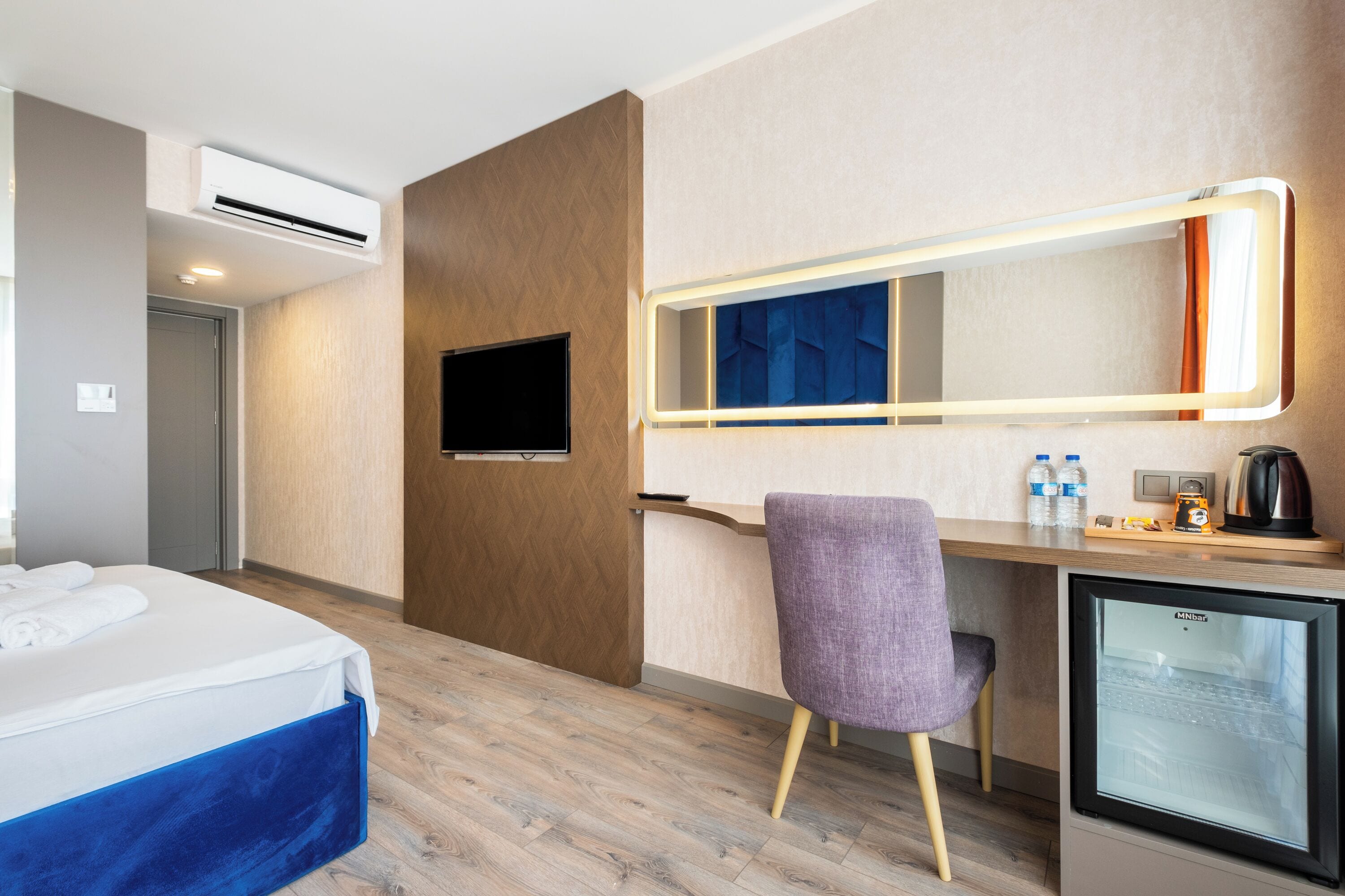 A hotel room with a bed, a desk with a refrigerator, a chair, a television, and a mirror with lights.