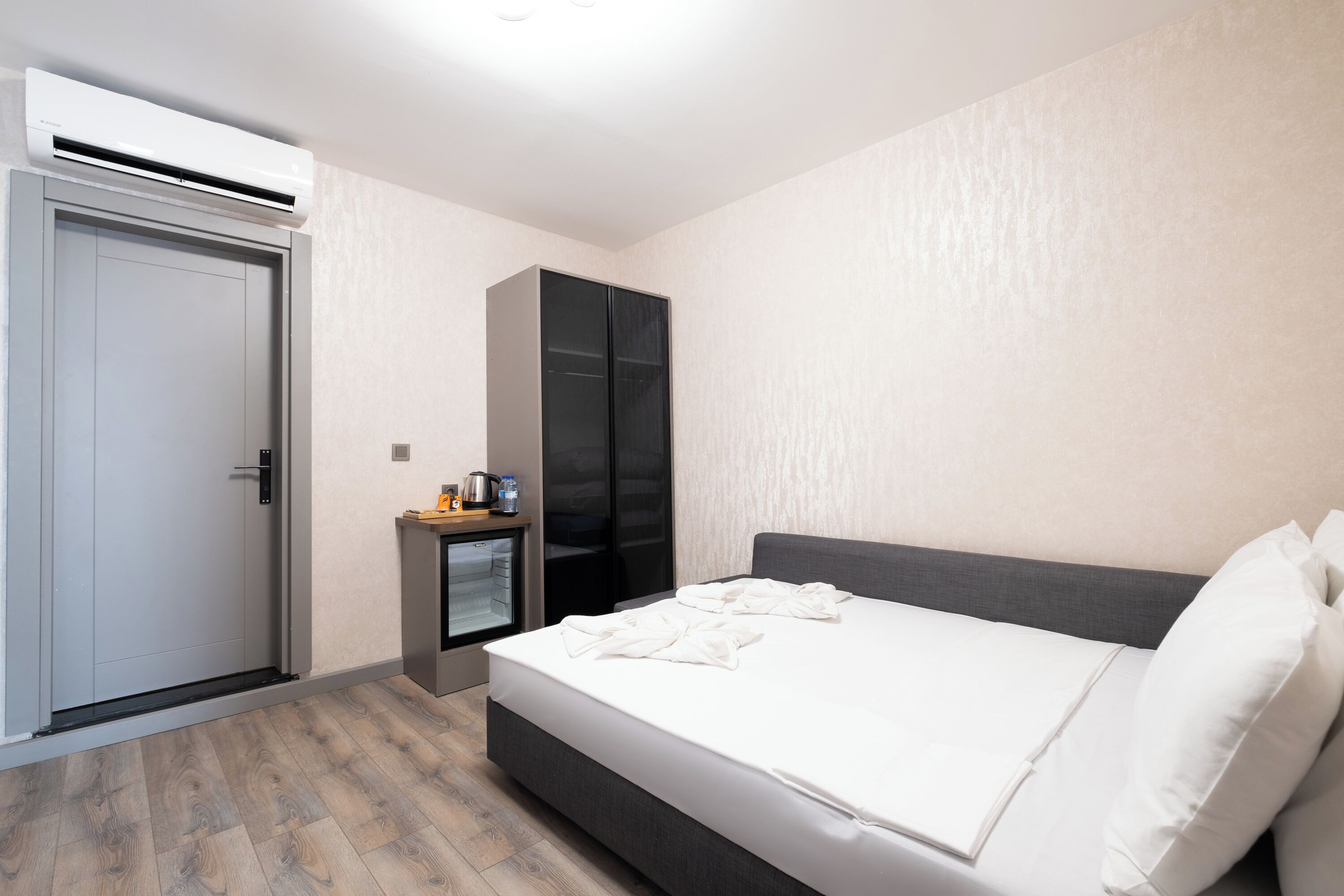 A modern hotel room with a bed, a wardrobe, a small desk with a lamp, and an air conditioning unit.