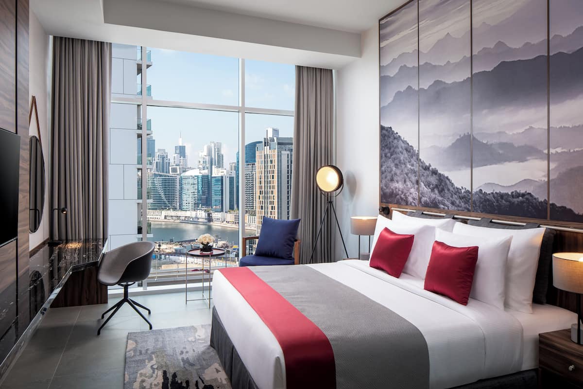 A hotel room with a large window offering a city view, a bed with red and white bedding, a desk with a chair, and a mounted TV.