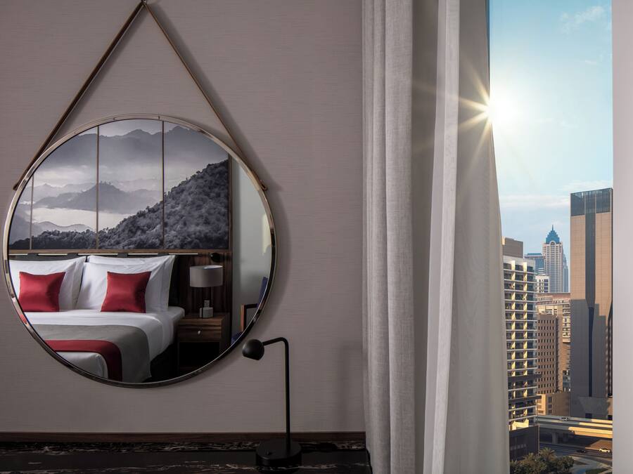A hotel room with a large round mirror reflecting a bed and a mountainous landscape. A window with a view of city buildings and a sun is visible.