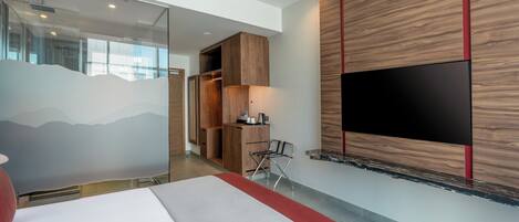 A modern hotel room with a large bed, a flat-screen TV mounted on the wall, a wooden wardrobe, and a small desk with a chair.
