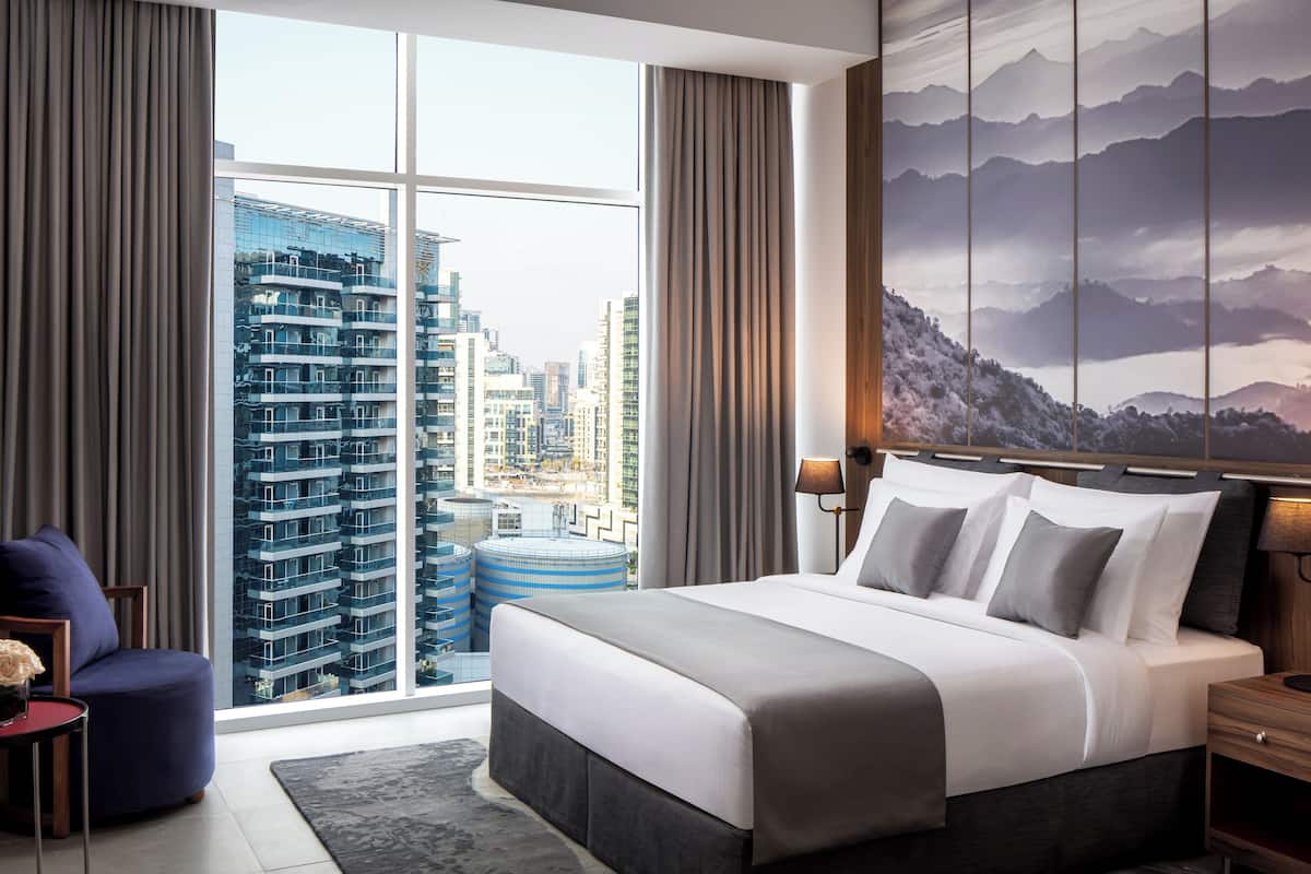 A modern hotel room with a large bed, a view of a cityscape through floor-to-ceiling windows, and a mountain landscape mural on the wall.