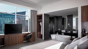 A modern hotel room with a large bed, a desk with a flat-screen TV, a walk-in closet, a bathroom with a bathtub, and a city view. - Grand Millennium Business Bay (Dubai)
