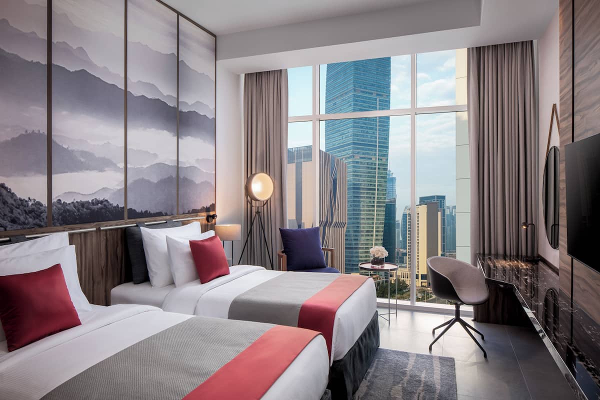 A hotel room with two beds, a TV, a desk, and a view of a cityscape.