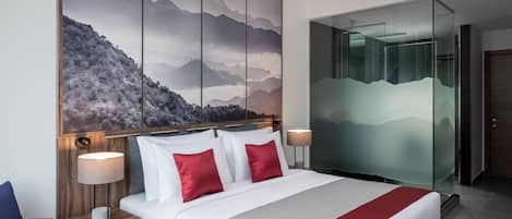 A hotel room with a large bed, two red pillows, a white comforter, a wooden nightstand with a lamp, and a large wall painting of mountains.