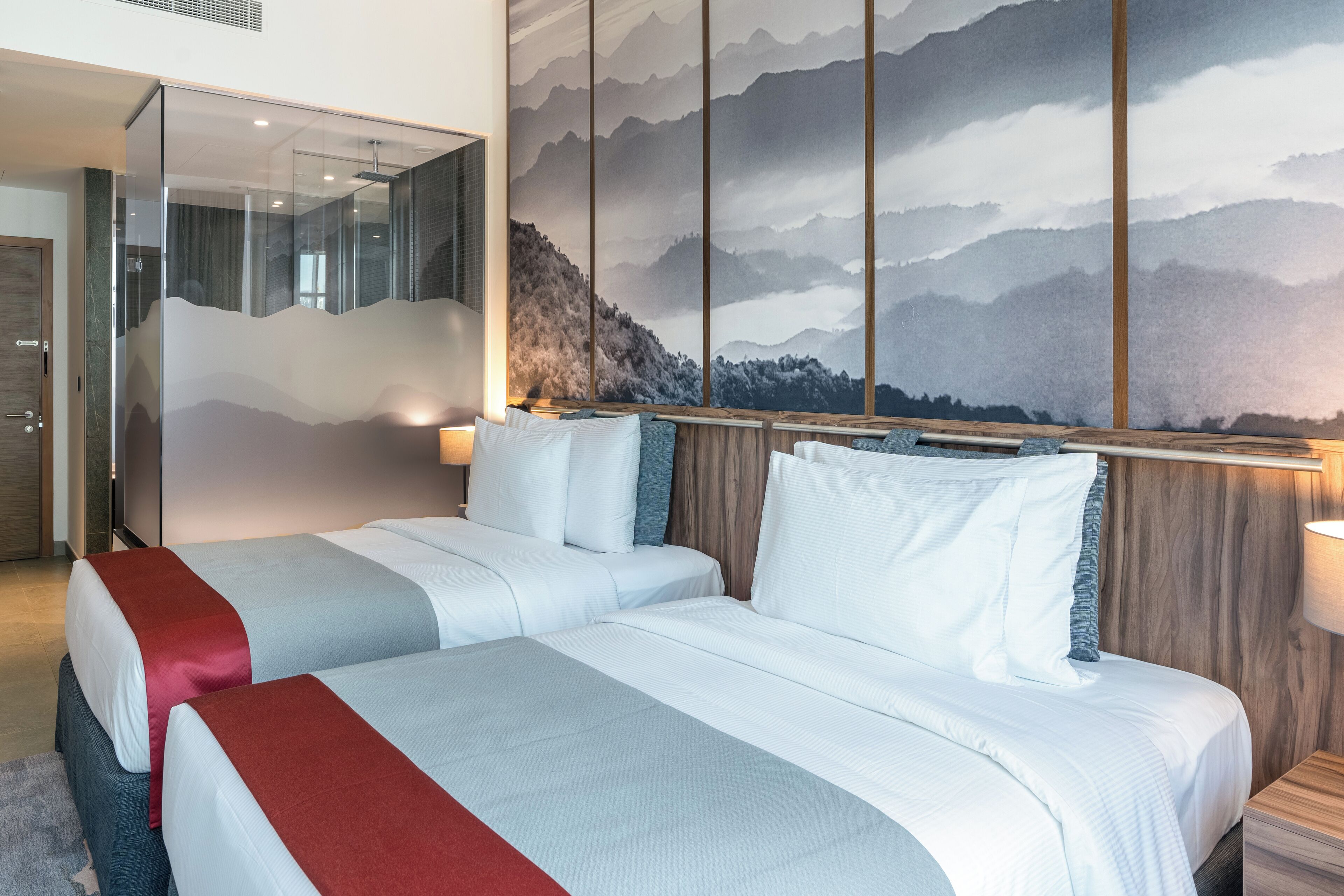 A hotel room with two beds, a large wall mural of mountains, and a glass partition.