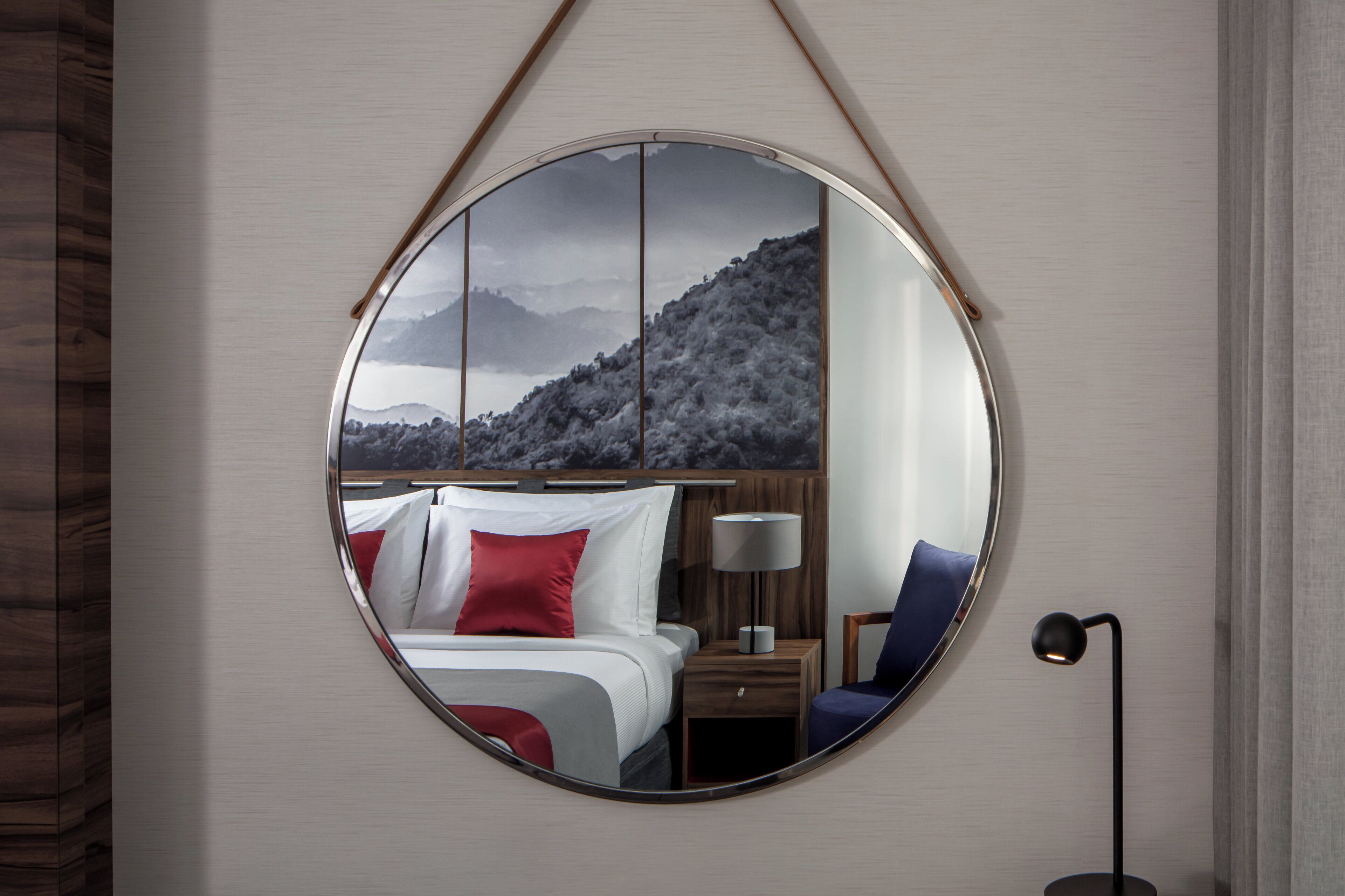 Ein Hotelzimmer mit Bett, Nachttisch, Lampe, Stuhl und einem großen runden Spiegel, der eine Berglandschaft reflektiert.
