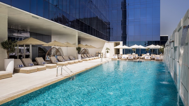 A swimming pool with lounge chairs and umbrellas, surrounded by a modern building with reflective glass windows. - Grand Millennium Business Bay (Dubai)