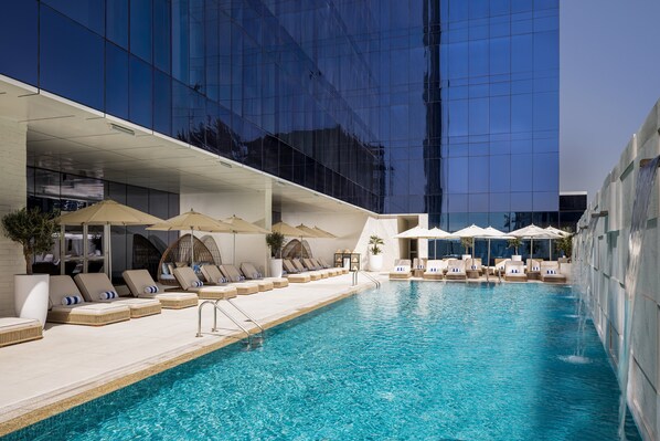 Piscina con sillas de descanso y sombrillas, rodeada por un edificio moderno con ventanas de vidrio reflectante.