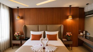 Une chambre d’hôtel avec un grand lit, deux tables de chevet, une petite table, un bureau avec une bouilloire et une fenêtre avec des stores.