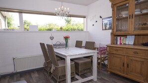Een eetgedeelte met een witte tafel en rieten stoelen, een houten kast met glazen deuren en uitzicht op groen.
