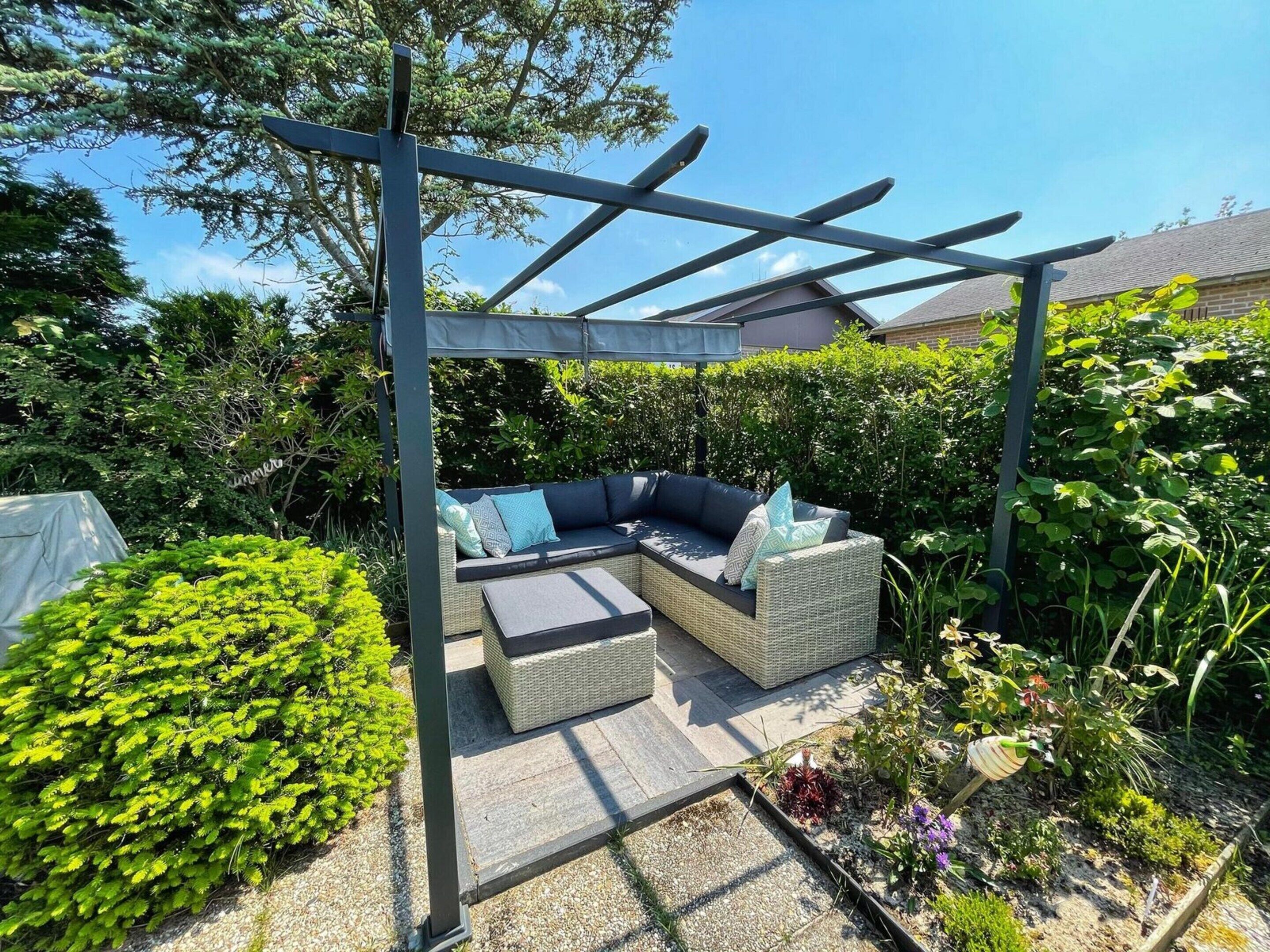 Zona salotto esterna coperta con divano in vimini e cuscini, immersa nel verde e caratterizzata da una pergola.