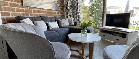 A modern living room with a grey sofa, two grey armchairs, a white coffee table, and a flat-screen TV on a white cabinet. A painting of a landscape is on the brick wall, and a window with patterned curtains is visible.