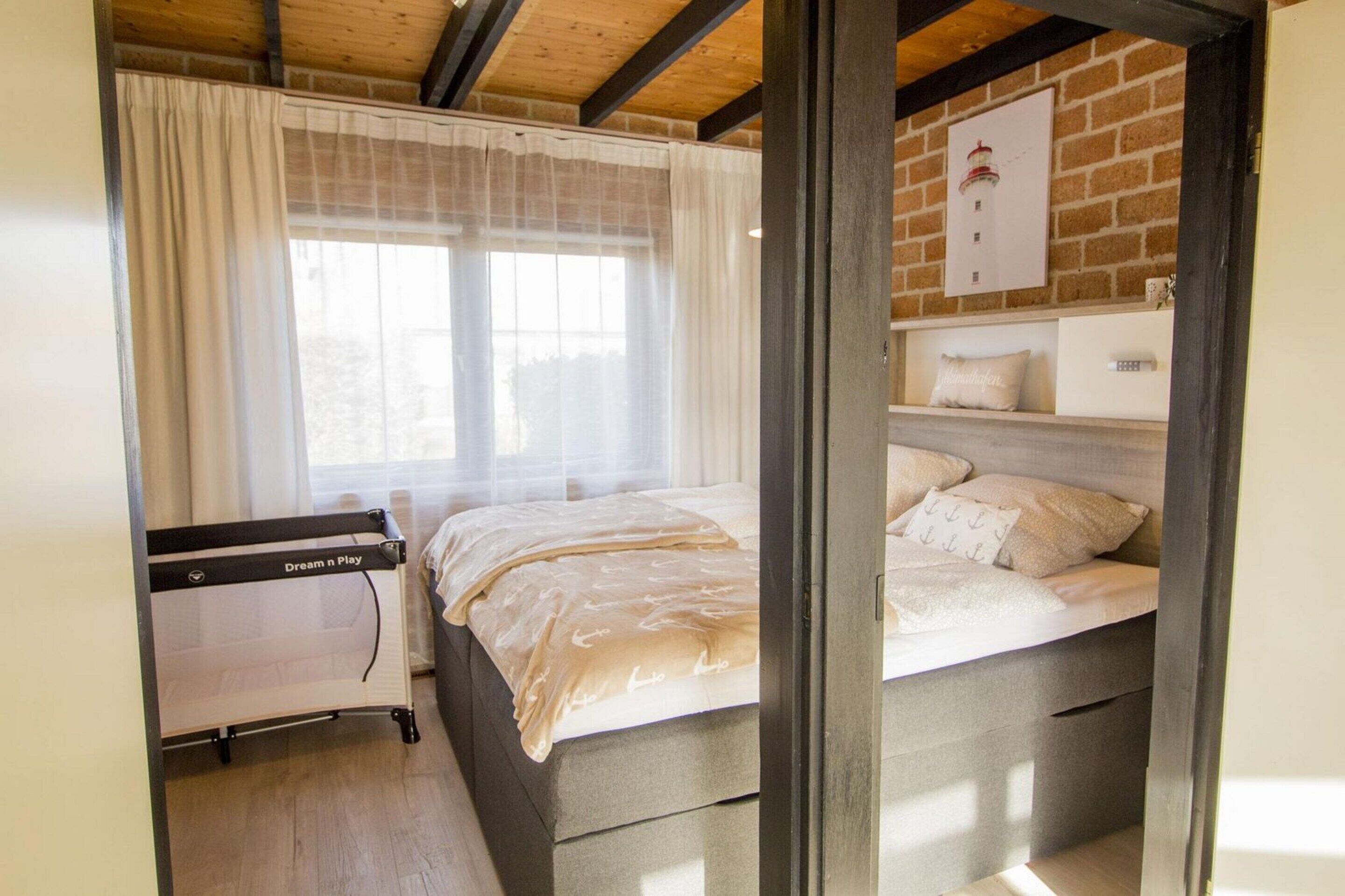 A bedroom with a bed, a bedside table, a nightstand, a window with curtains, a brick wall, and a wooden ceiling.