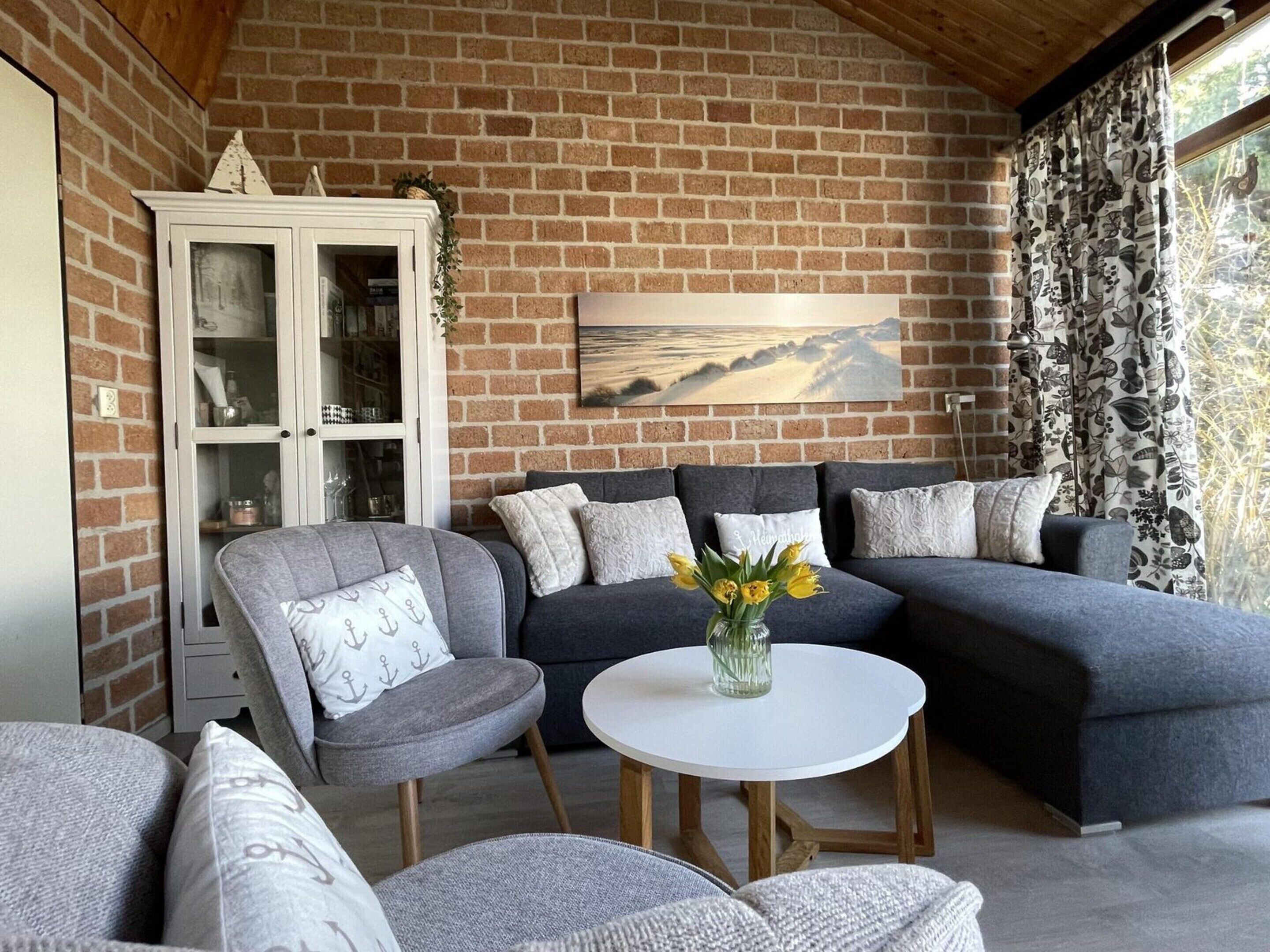 Een gezellige woonkamer met een bakstenen muur, een grijze hoekbank, een wit kastje en een schilderij van een landschap.