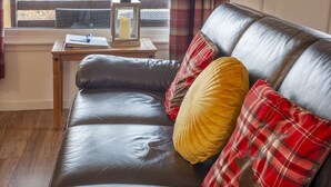 A leather sofa with red and yellow plaid cushions, a wooden side table with a lantern, and a window with a view of a wooden deck. - Birch Tree Lodge - sleeps 4 guests in 2 bedrooms (Appin)