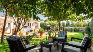 Ein Außenbereich mit Terrassenausstattung, Korbmöbeln, einem Tisch mit Flasche und Gläsern sowie Blick auf ein Haus mit Terrasse und Pflanzen.