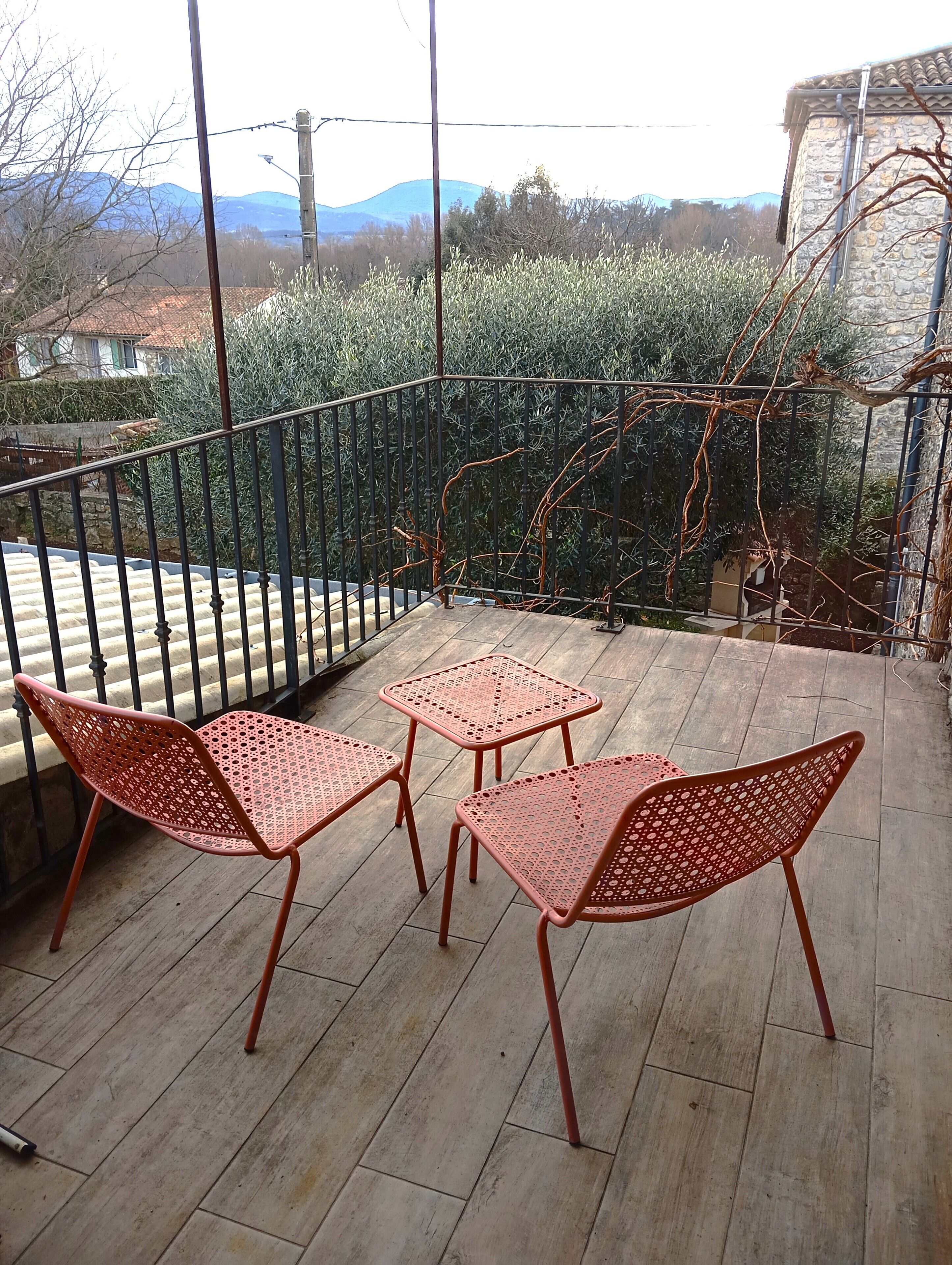 A balcony with three red metal chairs and a small table, overlooking a garden with a stone wall and a view of distant mountains.