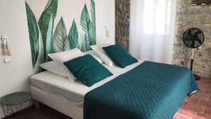 Una cama con ropa blanca y almohadas verde azuladas, un edredón verde azulado, una lámpara de pared, una pequeña mesa redonda, un ventilador y una pared de piedra.