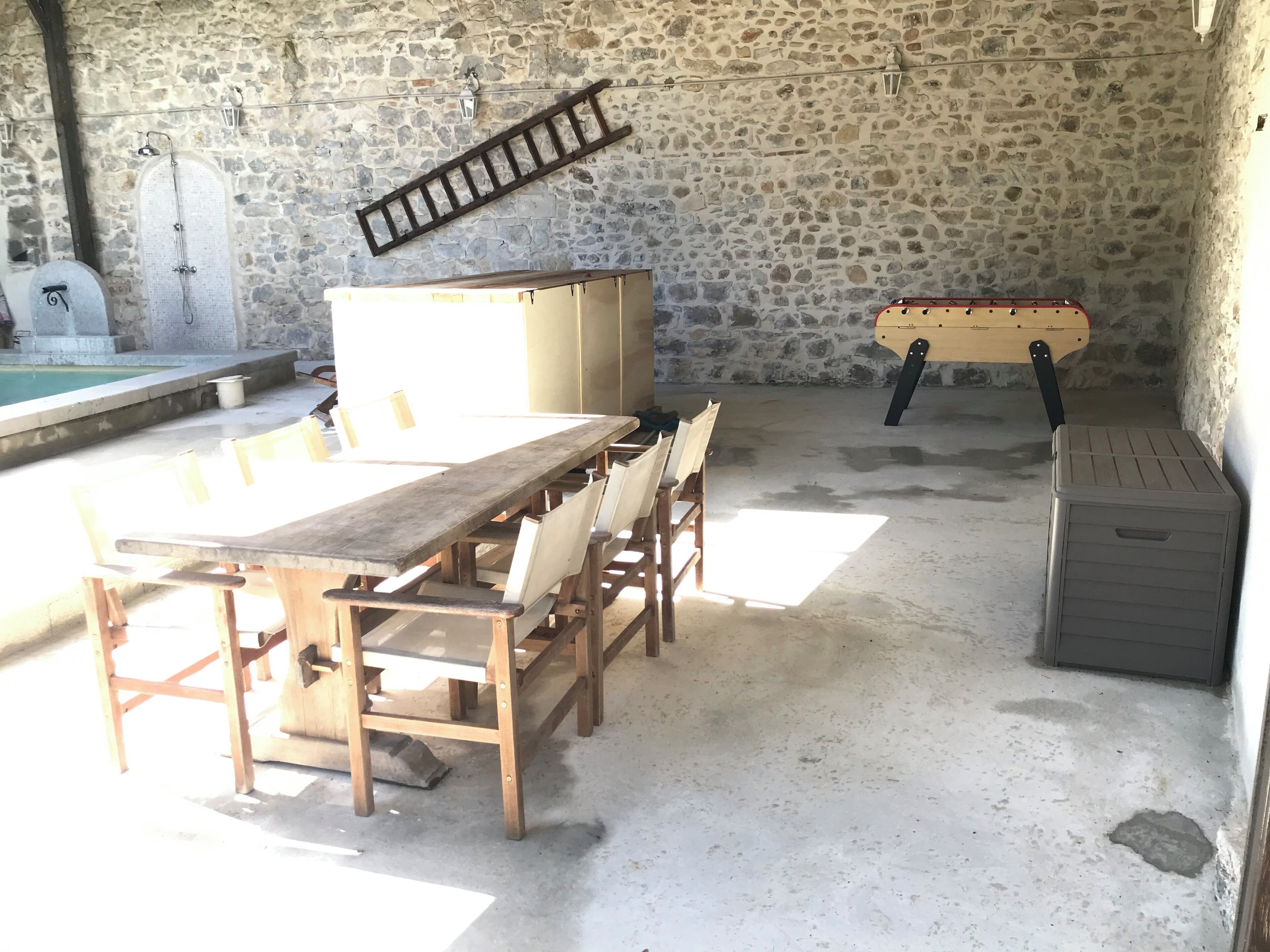 A stone-walled room with a long wooden table and chairs, a small pool, and a wooden bench.